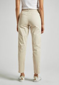 Chino in cotone beige con design a gamba dritta, dotati di tasche posteriori e una texture liscia, abbinati a sneakers argentate e trasparenti.