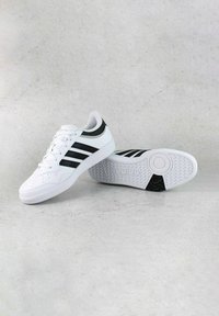 Scarpe da ginnastica bianche con strisce nere, punta perforata, colletto imbottito e suola in gomma testurizzata con un design circolare e logo.