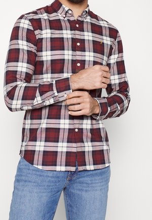 Mężczyzna poprawia rękaw czerwono-biało-czarnej koszuli w kratę zapinanej na guziki, nosi niebieskie dżinsy, stoi na białym tle.