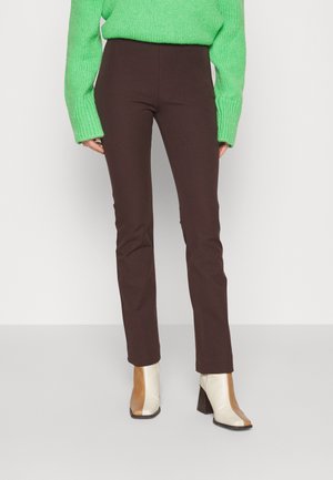 Persona con pantalones ajustados de color marrón oscuro, suéter de manga larga verde brillante y botines beige y marrones de dos tonos con tacón, de pie.