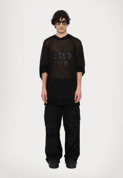 Schwarzes Mesh langärmliges Shirt mit "032c BERLIN" Druck, kombiniert mit lockeren schwarzen Cargo-Hosen. Das Modell trägt dunkle Sonnenbrillen und schwarze Schuhe.