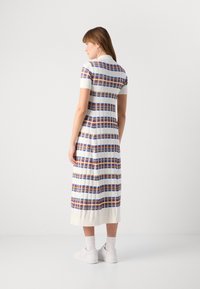 Γυναίκα που στέκεται με το πρόσωπο στραμμένο προς την αντίθετη κατεύθυνση, φορώντας ένα λευκό φόρεμα μέσου μήκους με μπλε, πορτοκαλί και καφέ οριζόντιες καρώ ρίγες και λευκά αθλητικά παπούτσια.