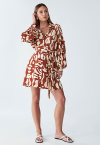 Brązowo-kremowa sukienka z abstrakcyjnym wzorem, z długimi rękawami, zapinana na guziki z przodu, z paskiem w talii. Lekki materiał, długość do połowy uda.