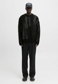 Eine Person trägt einen schwarz-grauen gemusterten Pullover, lockere schwarze Hosen, dunkle Schuhe und eine schwarze Mütze mit weißem Text und steht vor einem weißen Hintergrund.