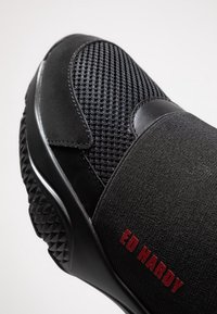 Schwarzer Sportschuh mit einem Obermaterial aus Netzgewebe, glatten Akzenten, elastischen Riemen, strukturiertem Sohlen und rotem "ED HARDY" Branding.