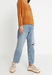 Przesadny, prążkowany pomarańczowy sweter zestawiony z jasnoniebieskimi, podartymi, luźnymi dżinsami oraz beżowymi trampkami na platformie z brązowymi podeszwami.