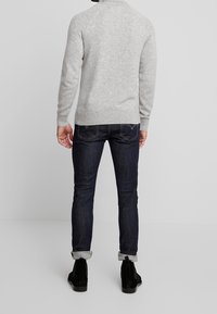 Homme portant un pull gris clair, un jean bleu foncé retroussé et des bottes noires, debout contre un fond clair uni, dos tourné vers le spectateur.
