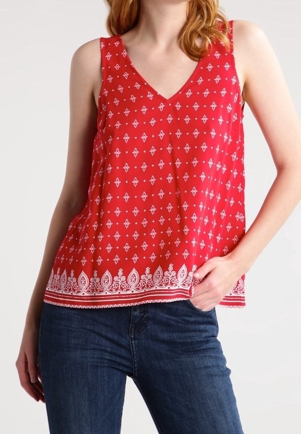 Femme portant un haut rouge sans manches avec des motifs géométriques et floraux blancs, associé à un jean bleu foncé, posant avec une main dans la poche.