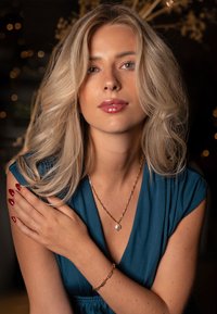 Blonde Frau in einem türkisfarbenen Kleid, mit einer goldenen Kettenhalskette mit einer Perle und einem goldenen Armband, die samtartige Haut und rote Nägel zeigt.