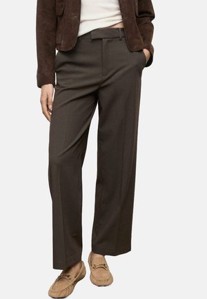 Vrouw draagt donkerbruine broek met wijde pijpen, beige loafers, een wit topje en een donkerbruine suède jas met één hand in de zak.