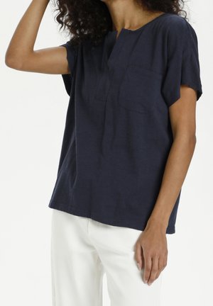 Granatowy, krótki rękaw, top z okrągłym dekoltem, detalem w postaci kieszeni z przodu, luźnym krojem i miękką fakturą materiału. Noszony z białymi spodniami.