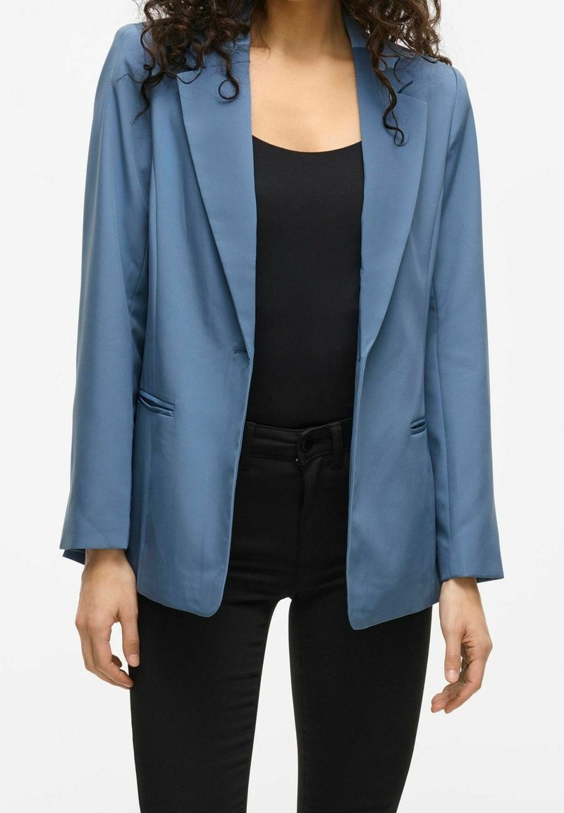 Femme portant un blazer bleu clair sur un haut noir et un pantalon noir, debout devant un fond blanc.