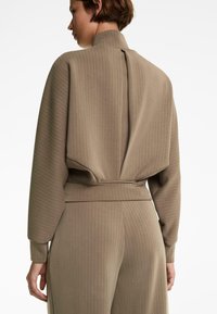 Donna con indosso una blusa beige a righe con colletto alto e pantaloni larghi abbinati, vista di spalle su uno sfondo bianco.
