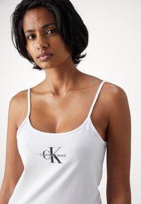 Hvit bomullstopp med tynne stropper, med et svart brodert "CK"-logo i midten. Glatt tekstur og slim fit-design.
