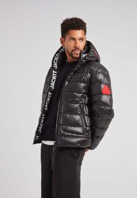 Giacca a vento nera in materiale lucido, design trapuntato, tasche con zip e cappuccio con branding bianco. Emblema rosso sul braccio sinistro.