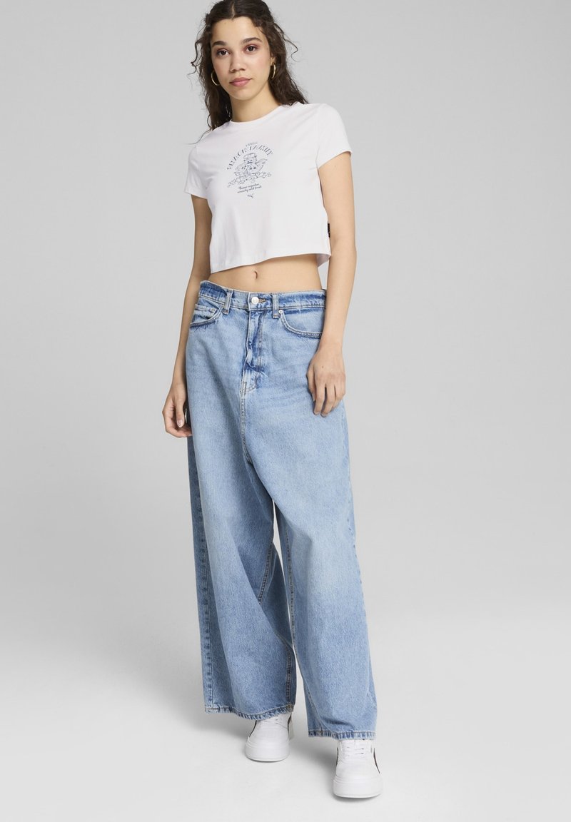 Hvid cropped t-shirt med grafisk print, parret med lyseblå vide ben jeans med høj talje og subtilt slid, båret med hvide sneakers.