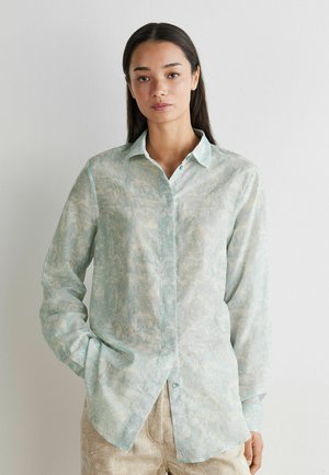 Young woman with long dark hair wears a pale blue patterned button-up shirt and beige pants, standing against a white wall.