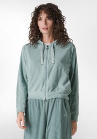 Sudadera con cremallera de pana en color verde azulado claro, con bolsillos frontales, capucha con cordón y puños acanalados, combinada con pantalones a juego.