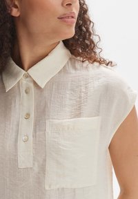 Cremfarbendes, ärmelloses Hemd aus einem weichen, strukturierten Stoff; verfügt über eine Knopfleiste und eine Brusttasche.