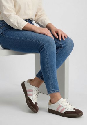 Osoba ubrana w niebieskie obcisłe dżinsy i białe trampki z różowymi paskami, siedząca na białej ławce na jednolitym tle.