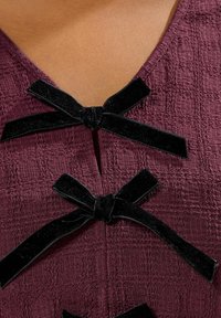 Primo piano di un indumento viola con una texture, dotato di due fiocchi in velluto nero legati al colletto.