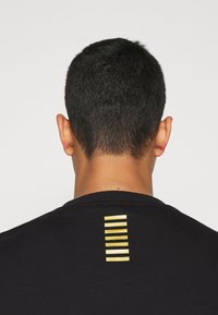 Svart t-shirt med rund krage och en vertikal rad av tre guldstrimmor på övre ryggen. Slät textur, enkel design.