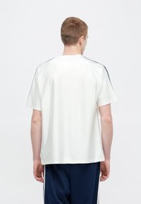 Biele tričko s krátkym rukávom a hladkou textúrou. Má raglanové rukávy a námornícke modré akcenty pozdĺž ramien. Voľný strih.