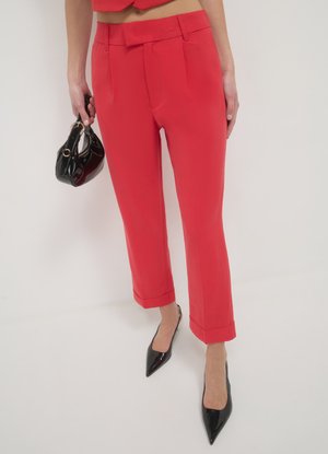 Pantalon rouge sur mesure avec taille haute et plis. Ourlet aux chevilles. Associé à des talons noirs à bouts pointus et un petit sac à main.