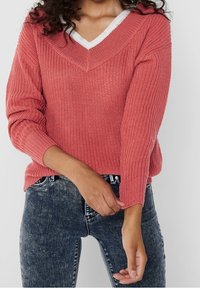 Koraalkleurige gebreide trui met een V-hals, geribbelde manchetten en zoom. Gedragen over een witte shirt, met een gestructureerde afwerking en een losse pasvorm.