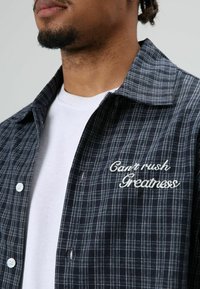 Overhemd met knopen in marineblauw met een geruit patroon. Heeft een witte borduring waarop links op de borst "Can’t rush Greatness" staat.
