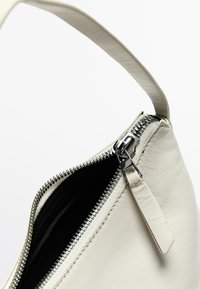 Borsa a spalla bianca in pelle con una cerniera metallica, caratterizzata da una superficie liscia e un design minimalista. Interno nero visibile attraverso l'apertura.