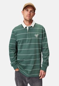 Grünes Langarm-Polo mit weißen horizontalen Streifen, ausgestattet mit einem weißen Kragen und besticktem Logo. Lässige Passform mit gerippten Bündchen.