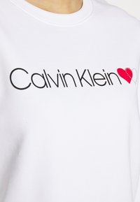 Felpa bianca con logo "Calvin Klein" in nero, decorata da un design a cuore rosso abbellito con piccole borchie per un tocco di dettaglio.