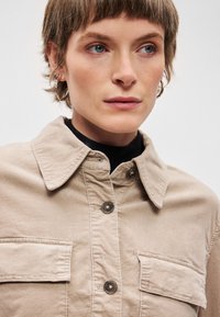 Veste en coton beige avec un col, deux poches à l'avant et des boutons-pression pour la fermeture. Texture subtile visible.