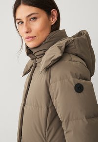 Khaki pufferjack met een hoge kraag, voorzien van een knoopaccent op de linkermouw en een gladde, gewatteerde textuur. Voorzien van een drukknopen sluiting aan de voorkant.