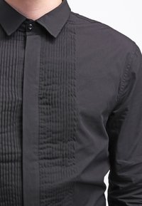 Mustat paita, jossa on napitettava kaulus, teksturoitu pleatitus edessä ja sileä kangas sivuilla. Ei näkyviä logoja tai kuvioita.