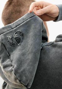 Rövid hajú személy, aki felhúzza egy szürke pulóver kapucniját, megmutatva a kopott hímzett logót a kapucnin.