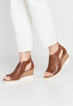 Brown perforated leather wedge sandals with open toe and cut-out design, featuring a textured cork base and white rubber sole.