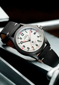 Montre Timex Expedition avec un boîtier en titane, un cadran blanc, un bracelet en cuir noir avec des coutures blanches et des accents rouges sur les index.