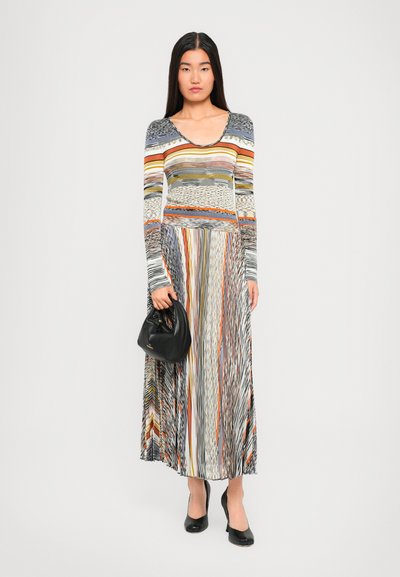 Donna che indossa un lungo vestito a strisce multicolori, con una borsa nera in mano, in piedi davanti a uno sfondo chiaro e uniforme.