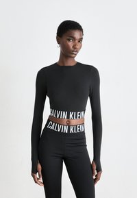 Dünne Person in schwarzem langärmligen Cropped-T-Shirt und hochtaillierten Leggings mit weißen "Calvin Klein"-Logo-Bändern, die vor einem schlichten weißen Hintergrund steht.