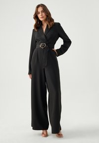 Zwarte jumpsuit met lange mouwen, uitgesneden kraag, een ceintuur met een ronde gouden gesp en wijde broekspijpen. Glad, lichtgewicht stof.