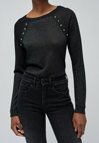 Frau in einem figurbetonten schwarzen Langarmtop mit dekorativen Knöpfen an den Schultern und hoch sitzenden schwarzen Jeans.