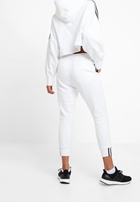 Bílá zkrácená mikina s kapucí, doplněná bílými zúženými tepláky s černými bočními pruhy a zúženými kotníky. Černé tenisky.