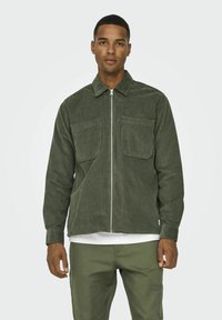 Veste en velours côtelé vert olive avec fermeture éclair à l'avant, col pointu et deux poches sur la poitrine. Manches longues avec poignets boutonnés et coupe décontractée.
