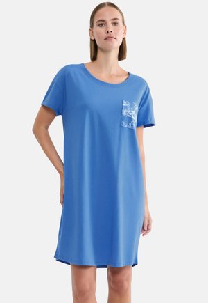 Kobieta ubrana w luźną, niebieską sukienkę z krótkim rękawem, z okrągłym dekoltem i wzorzystą kieszonką na piersi, stojąca na jednolitym tle.