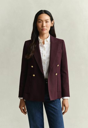 Vrouw met lang donker haar die een bordeaux dubbelgeknoopte blazer, wit overhemd en blauwe spijkerbroek draagt, staand tegen een egale lichte achtergrond.