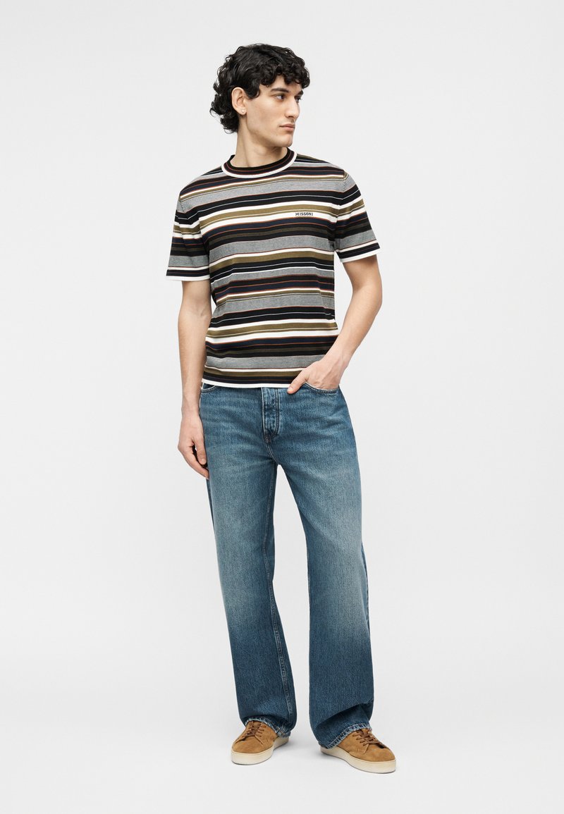 Man standing wearing a short-sleeve striped shirt, blue jeans, and brown sneakers, looking to his left with one hand in pocket.