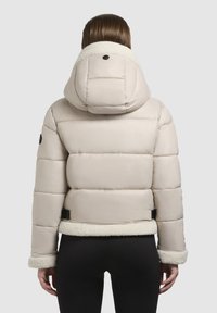 Naised, kes kannab heledat beežikat pufferjacketit, millel on kapuuts ja sherpa ääris, nähtuna tagant tasasel taustal.