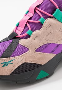 Gros plan sur une basket beige, violette et noire avec des lacets roses, des accents verts, des textures en mesh et en suède, et une semelle noire avec un détail vert.